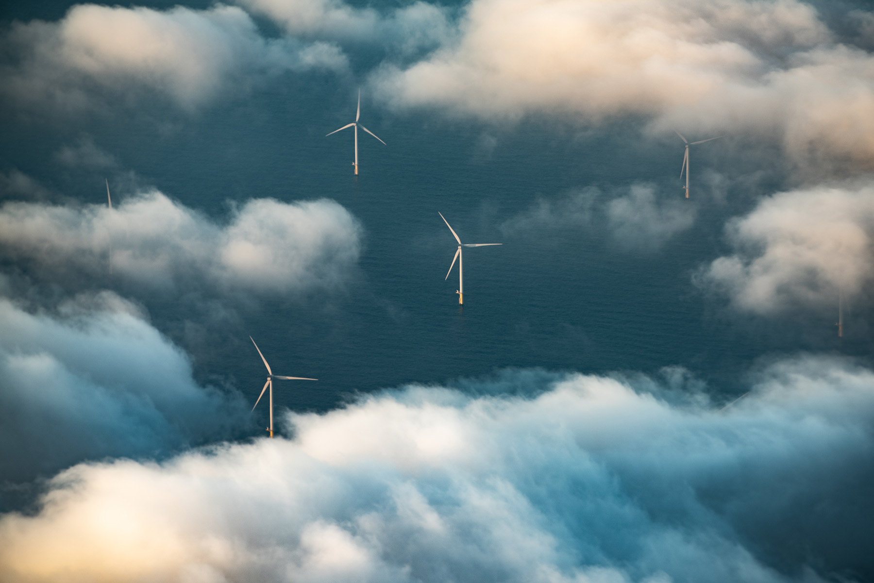 Five white wind turbines in an offshore wind farm are seen through white, fluffy clouds.