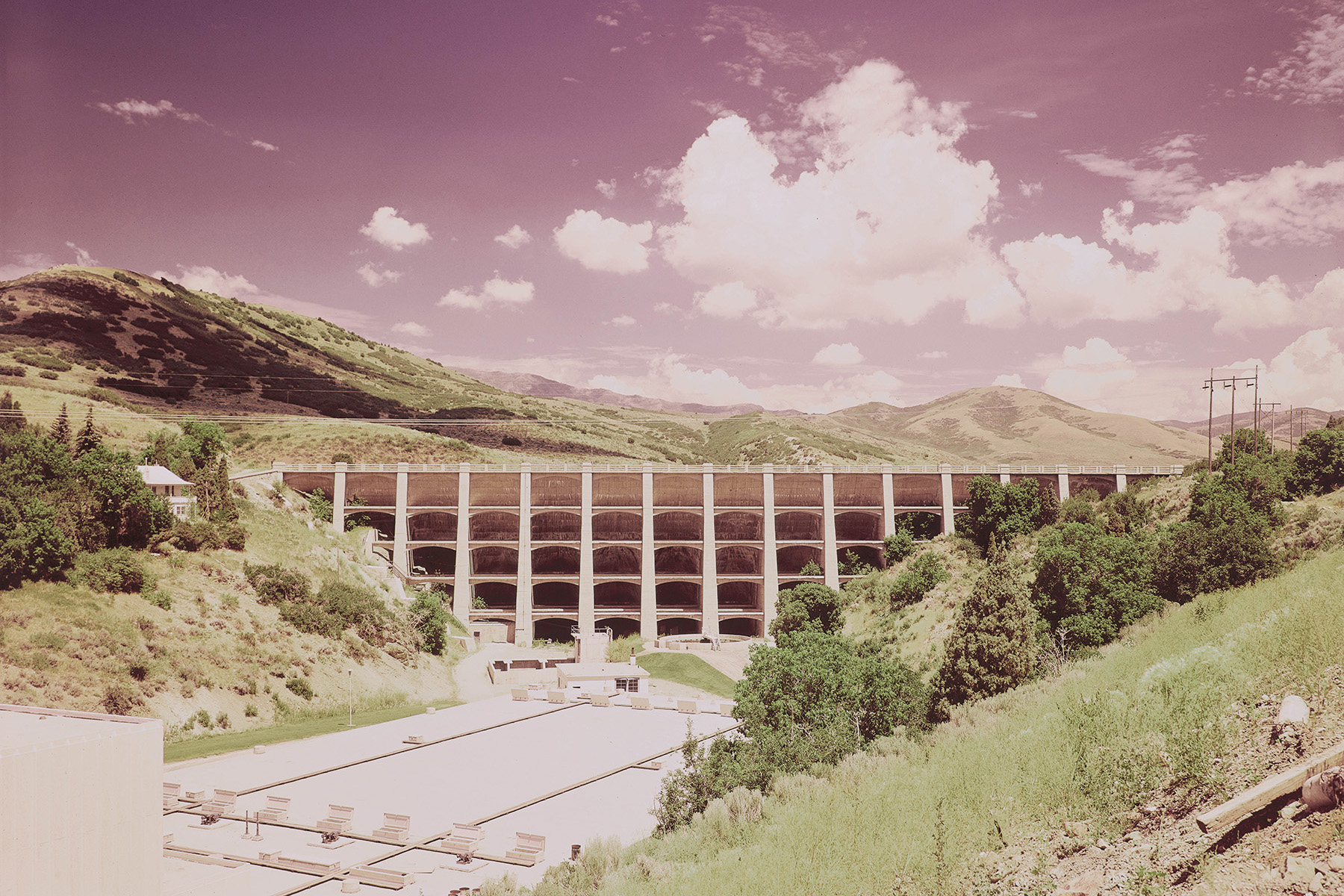 Photograph shows a multiple-arch dam set amidst trees and hills. 