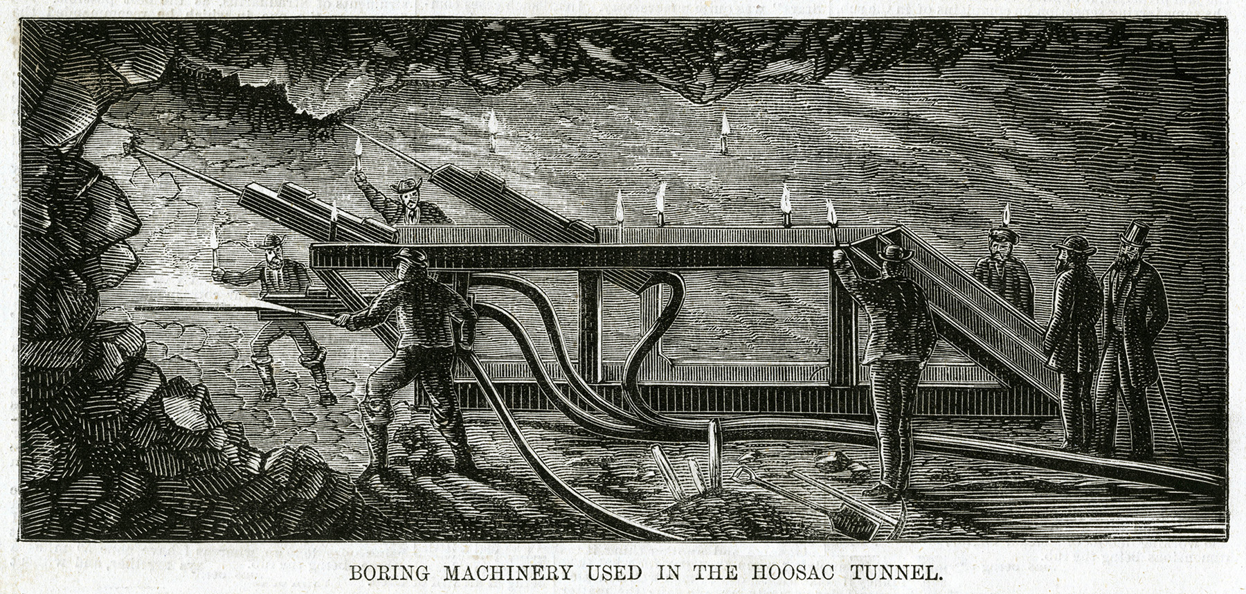 Men hold hoses that are used to blast away rock. Other men hold torches to light the explosive.