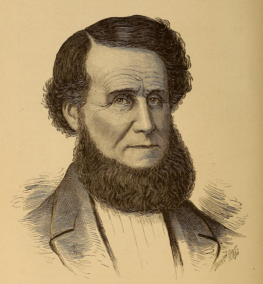 Image shows a main with curly hair and a full curly beard wearing a jacket and shirt.