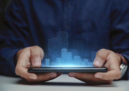 The image is a close-up of a pair of hands holding a device projecting digital images of buildings. 