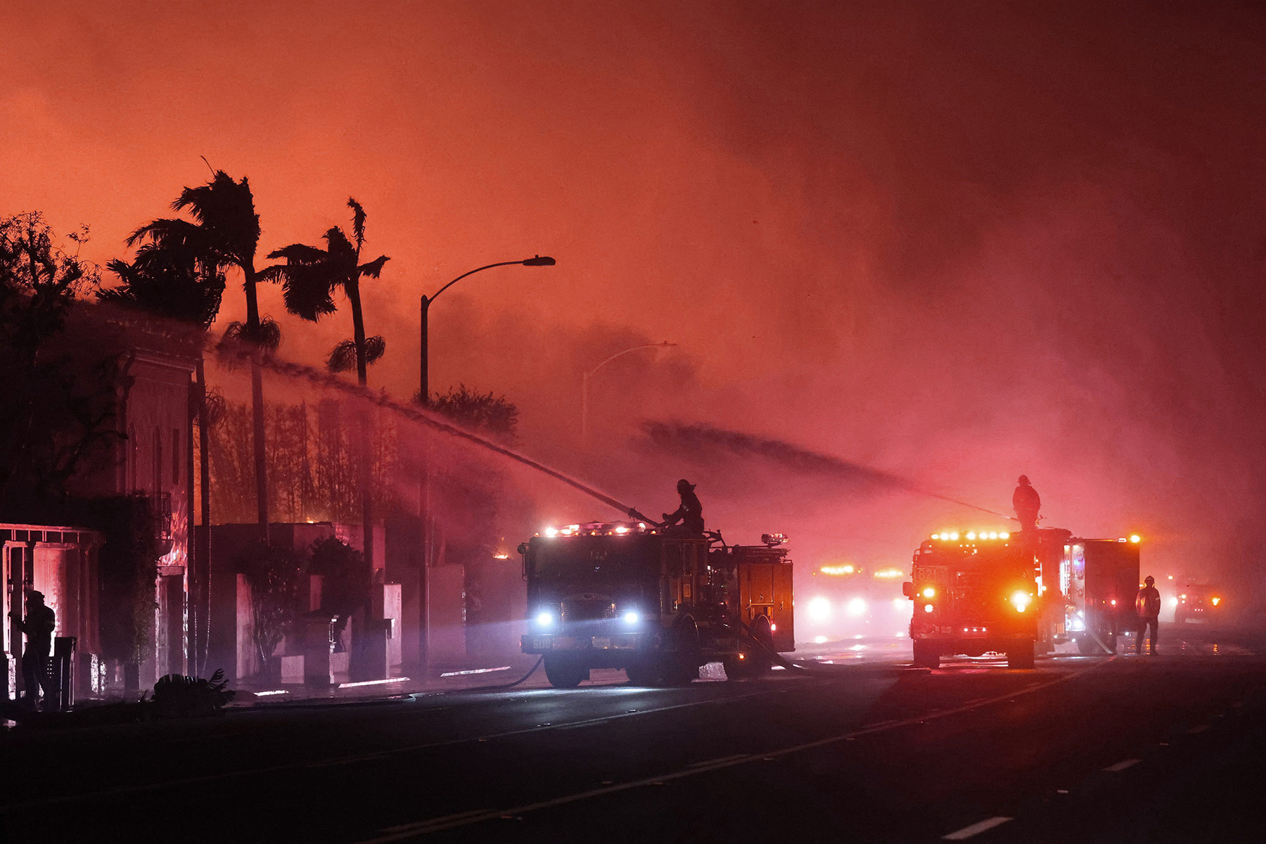 Two fire trucks spray water on burning buildings against a fire red sky.