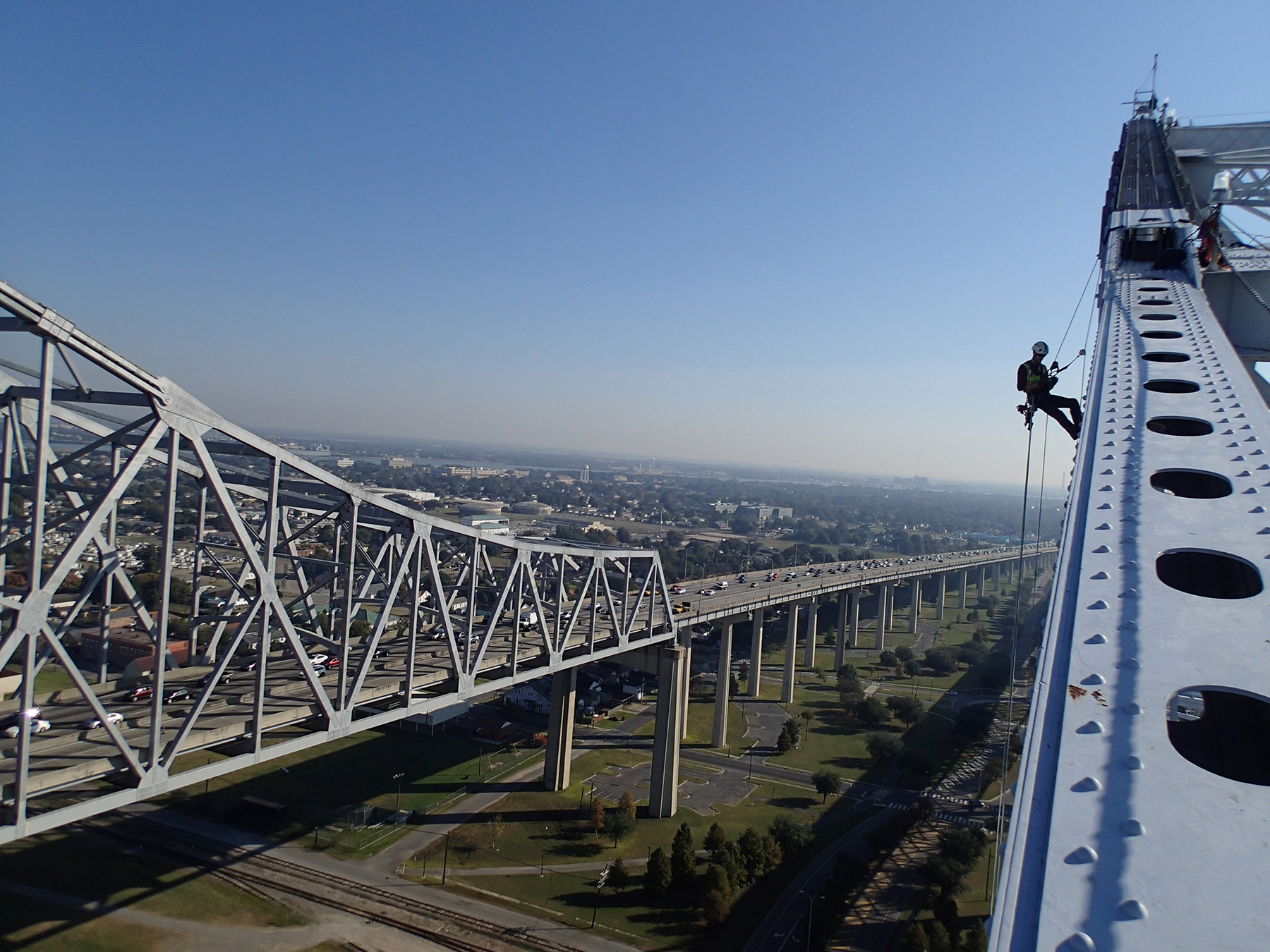 In the foreground, a person in full safety gear uses a rope system to inspect the top of a bridge. In the background is a multi-lane highway.