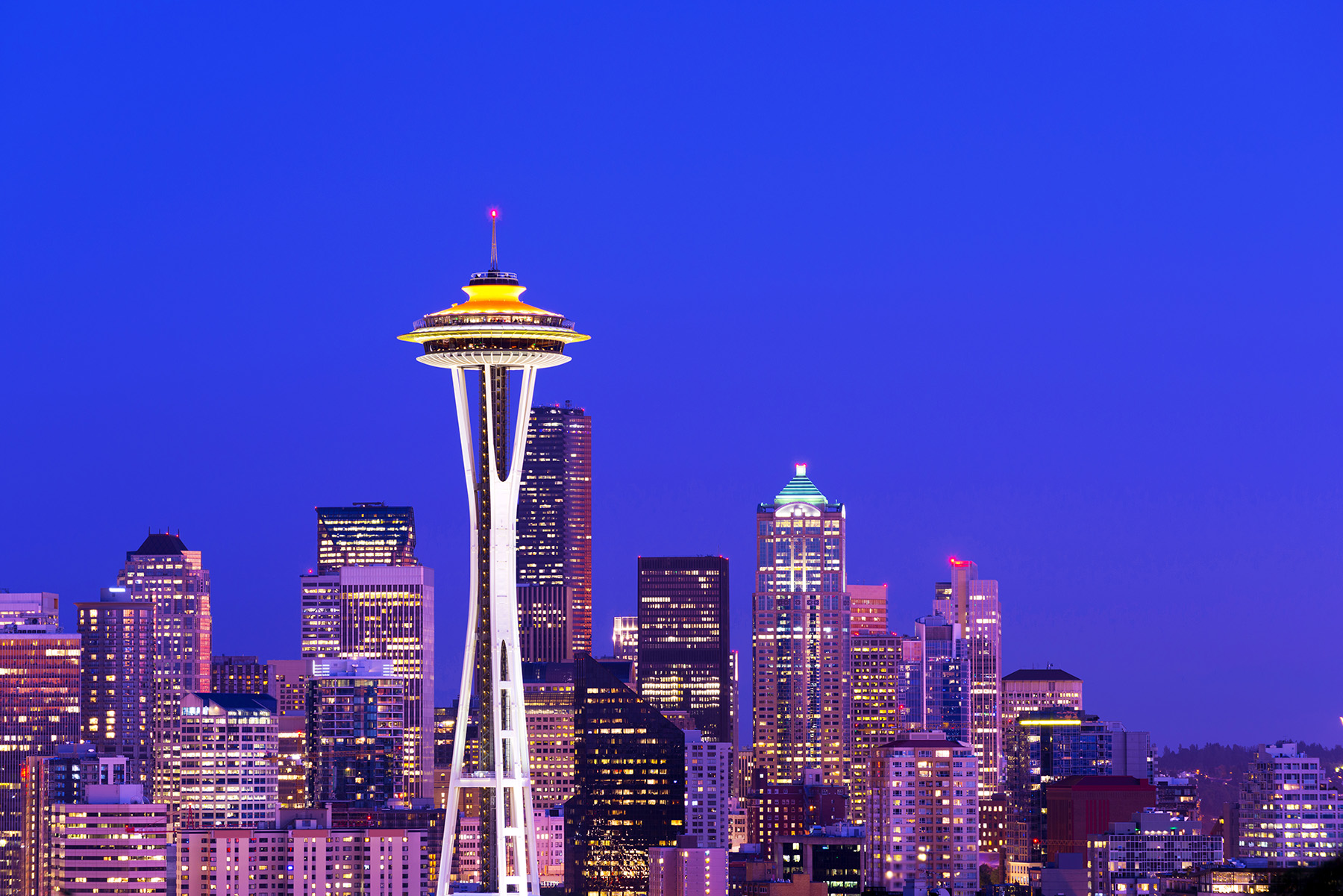 Photograph shows a structure with tall steel columns that taper in near the midsection with a saucer-like top lit up at night and surrounded by other buildings. The columns are connected to a central core. 